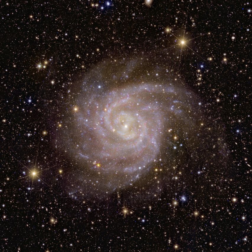 A big spiral galaxy is visible face-on in white/pink colours at the centre of this square astronomical image. The galaxy covers almost the entire image and appears whiter at its centre where more stars are located. 