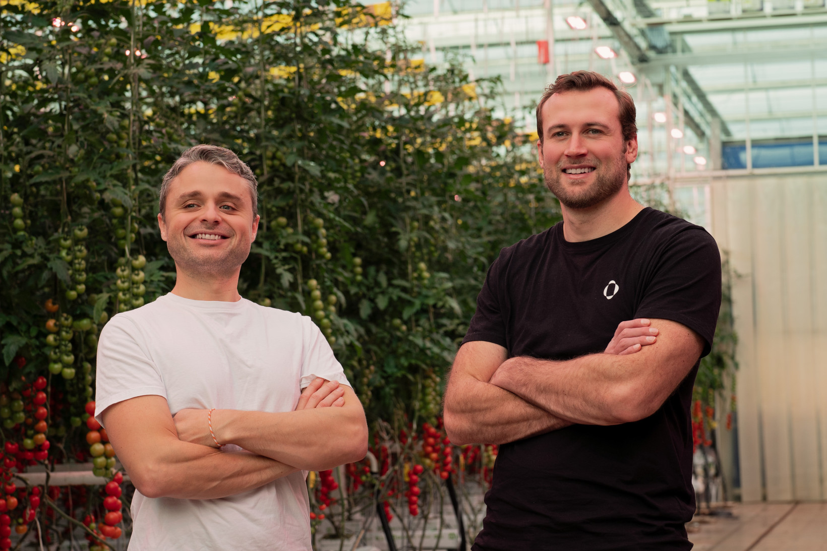 The founders of Source.ag standing in a greenhouse in front of plants, both are white men and one is wearing a white t-shirt and the other a black oned